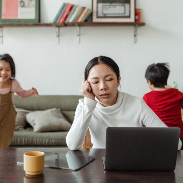 Tired-looking mother using a laptop while two children play in the background